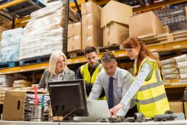 Gerentes de armazém e trabalhadores a trabalhar em mesa com computadores e ao fundo prateleiras de produtos