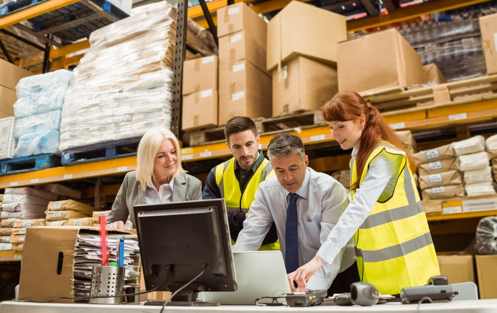 Gerentes de armazém e trabalhadores a trabalhar em mesa com computadores e ao fundo prateleiras de produtos